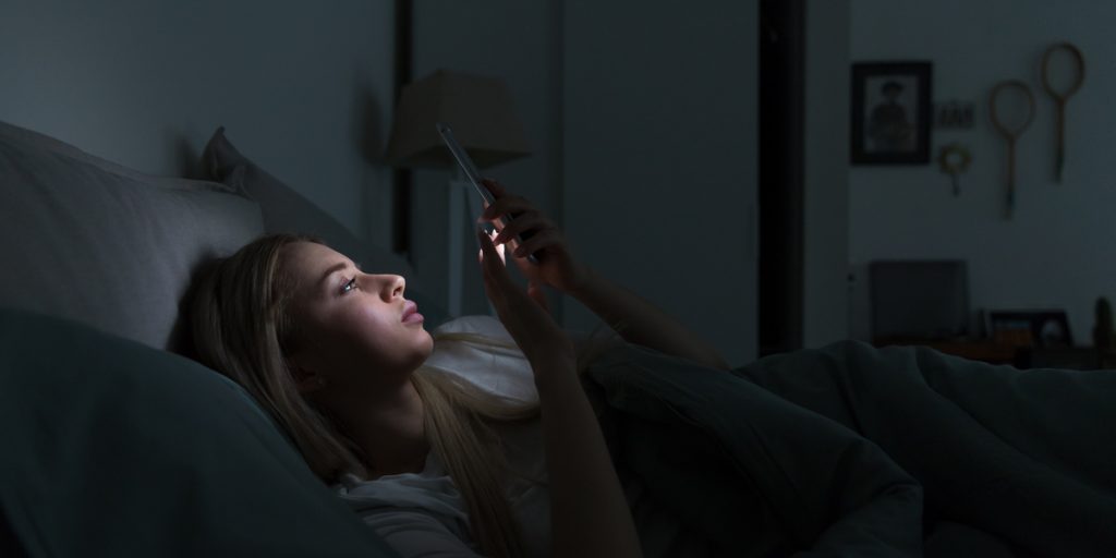 young woman laying in bed in a dark bedroom looking at cell phone