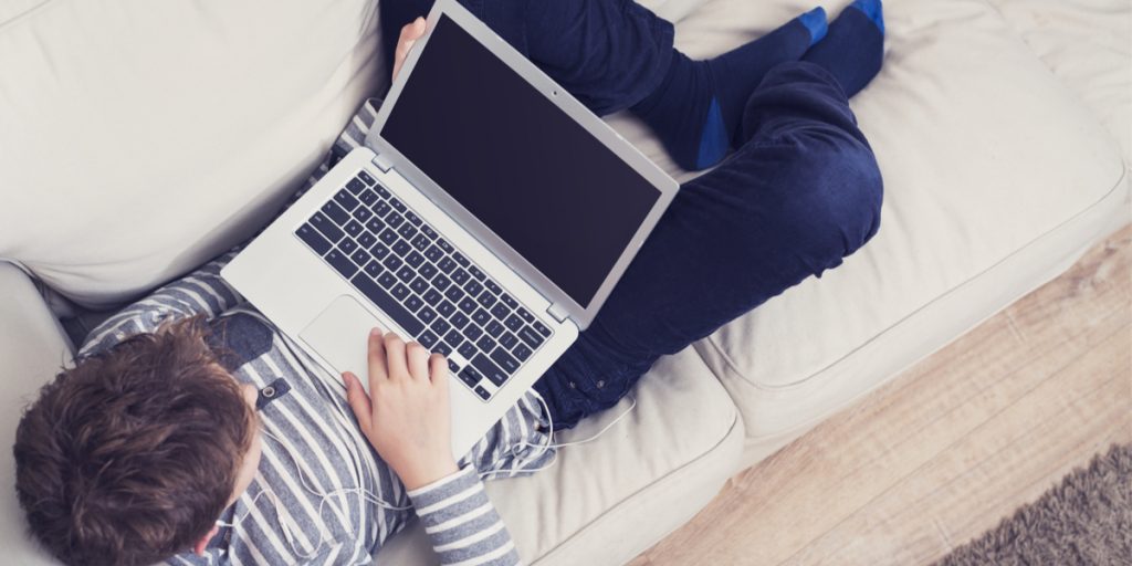 Adolescent boy laying on a couch with a laptop on his lap and earplugs in ears