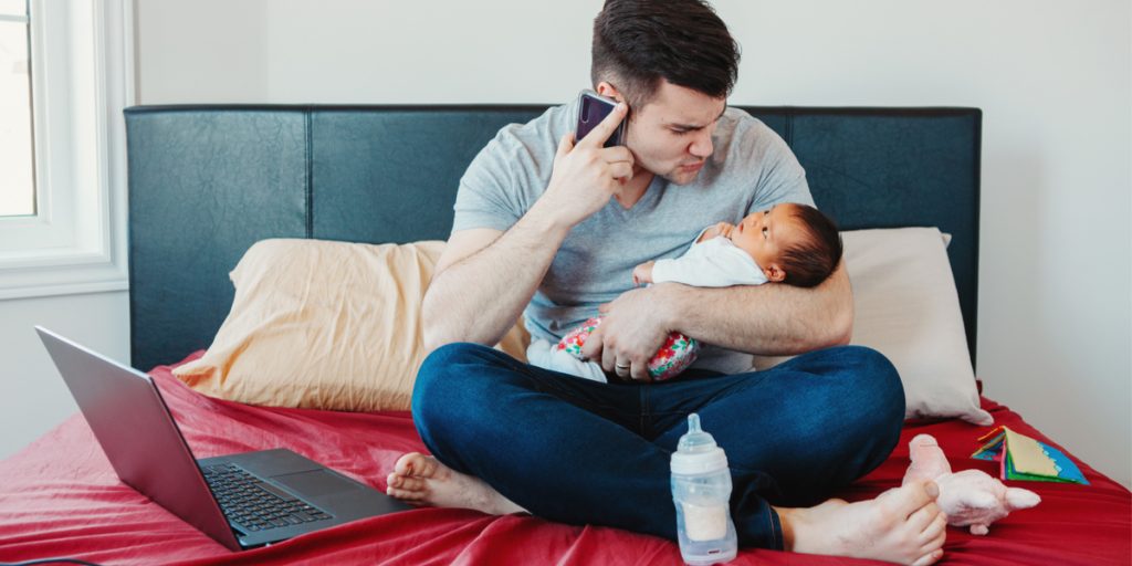 Man sitting on a bed holding a newborn baby in one arm and talking on a cell phone with the other hand and an open laptop on the bed next to the man