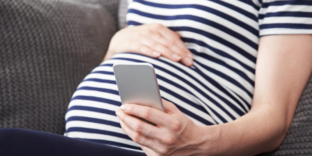 Close up of pregnant woman's belly while holding a cell phone in one hand next to her belly