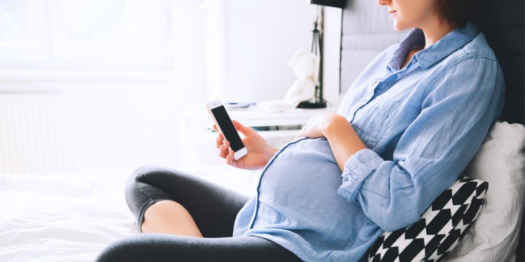 Close up of a pregnant woman sitting on a bed holding a cell phone in one hand close to her stomach and her other hand rested on her pregnant belly
