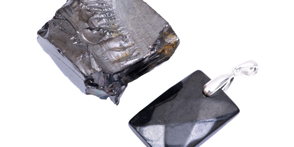 Rough Elite Shungite mineral next to smooth rectangular Elite Shungite on a pendant with a white background