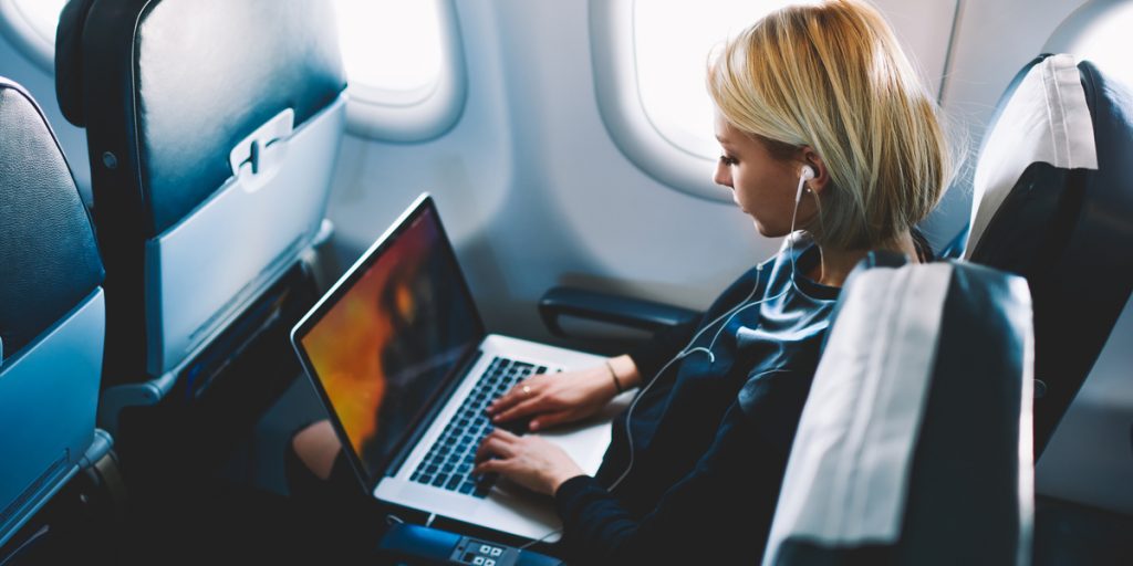 Woman sitting in an airplane with headphones in her ears working on a laptop on her lap