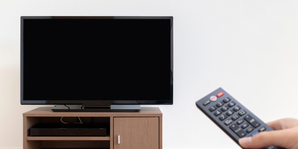 Off television on top of a TV stand with a hand outreached holding a remote control in the foreground