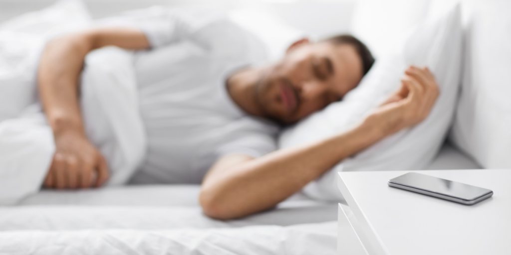 Man sleeping in a bed in the background with iPhone on a nightstand next to him in bed