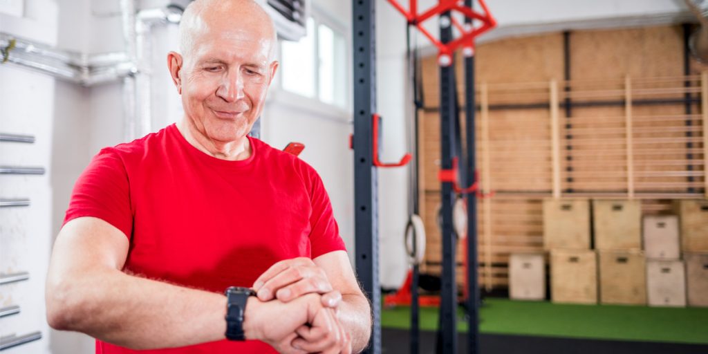 Older bald man wearing exercise close standing in front of a weight rack looking down at a fitness tracker watch on his wrist