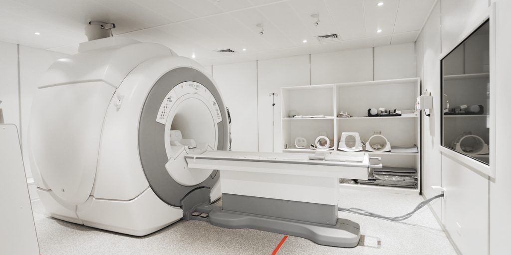 White MRI machine inside a white room with medical equipment on shelves and a look-through window on one wall inside the room
