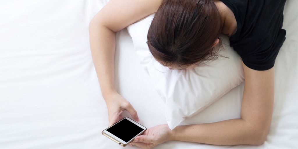 Top down view of woman laying on stomach in bed looking at an iPhone in her hands