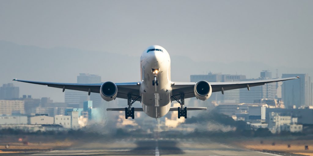 Front view of an airplane just at take off with the runway below the plane and a city in the background